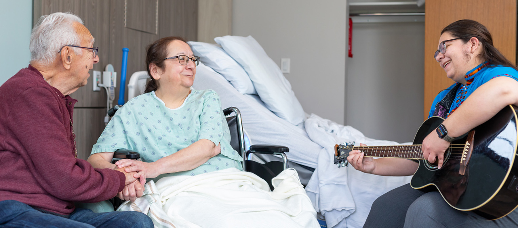 Wife and husband in Lightways hospice holding hands and listening with musician playing a guitar and singing for musical therapy.