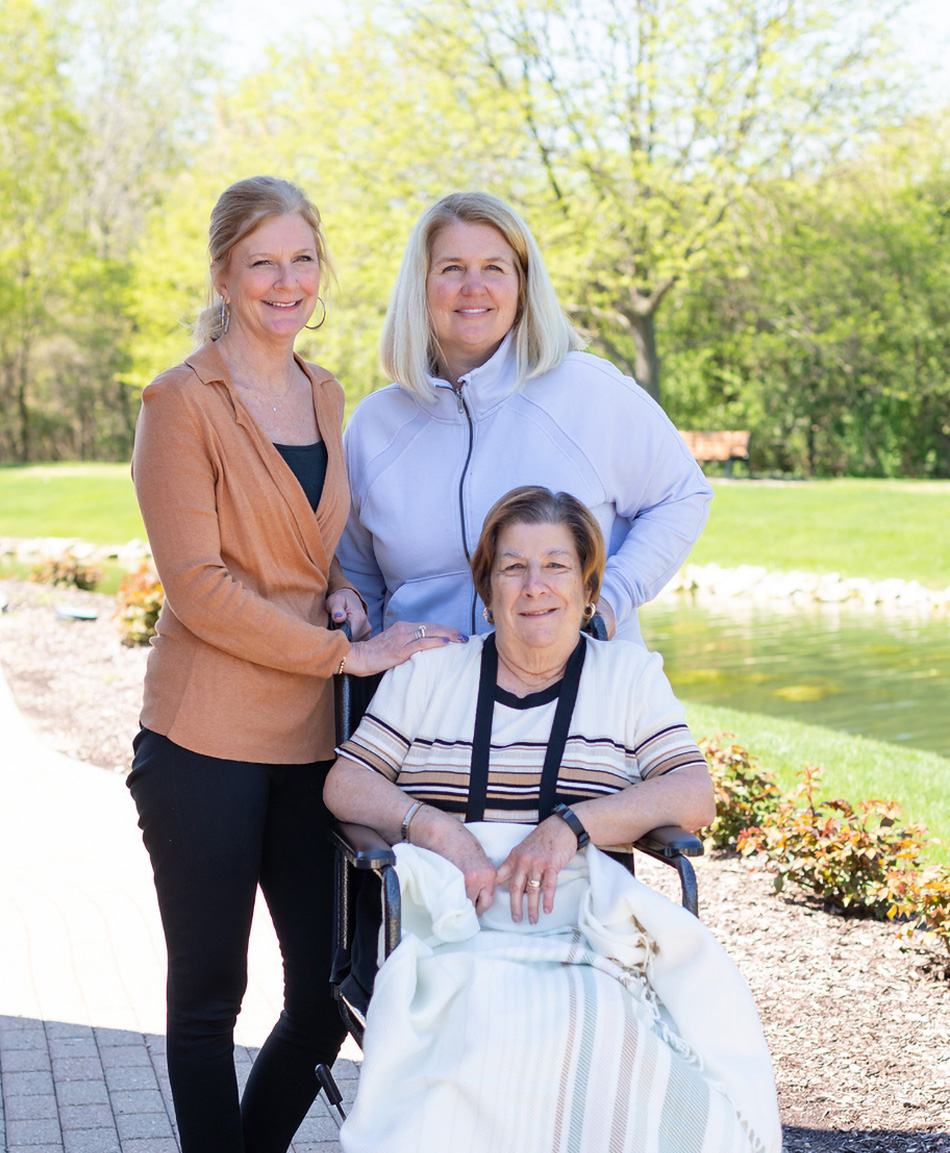 Mom and daughter stand with mother sitting in wheel chair outside by the lake at Lightways Joliet.